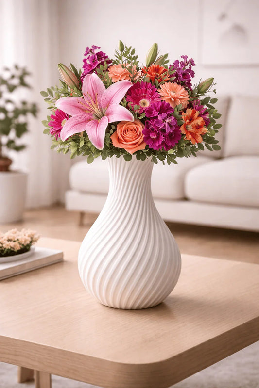 Jarrón blanco moderno con ramo de flores coloridas sobre mesa de madera en sala iluminada.
